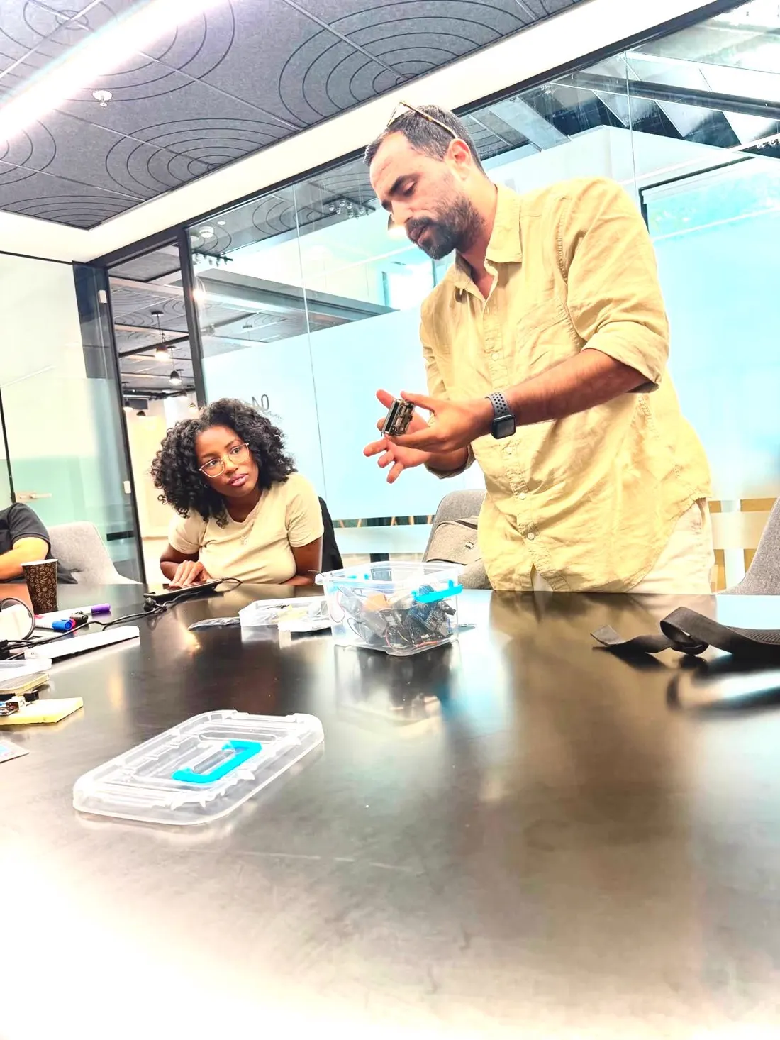 לידור עומד ליד שולחן הסדנה, מחזיק בקר ומסביר, ומשתתפת מתבוננת ברכיבים